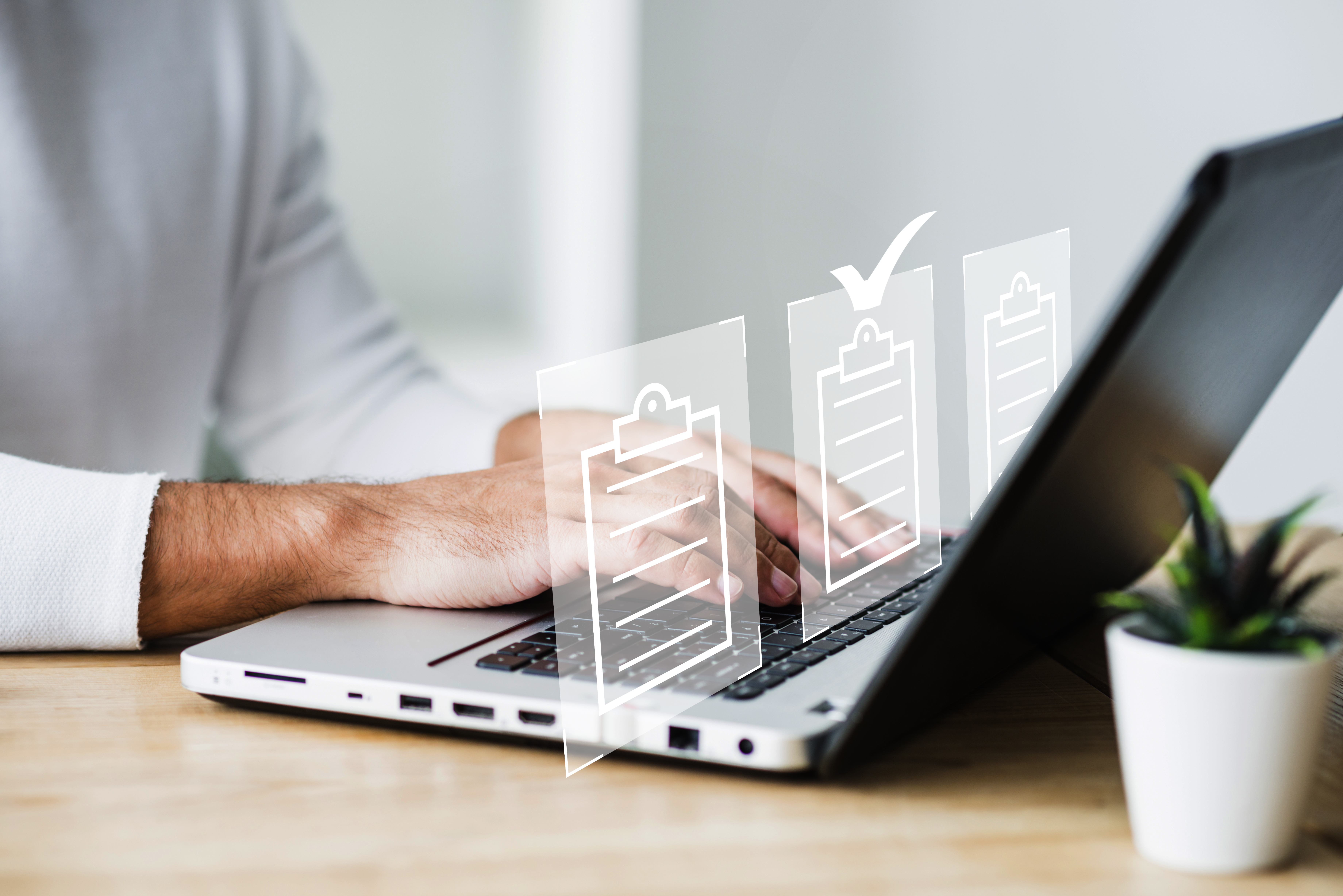 Profissional digitando em um notebook sobre uma mesa de madeira, com ícones transparentes de checklists sobrepostos à imagem e um símbolo de confirmação destacado, representando controle de processos, auditoria e conformidade. Ao lado do computador, há um pequeno vaso com planta, sugerindo ambiente de trabalho organizado.