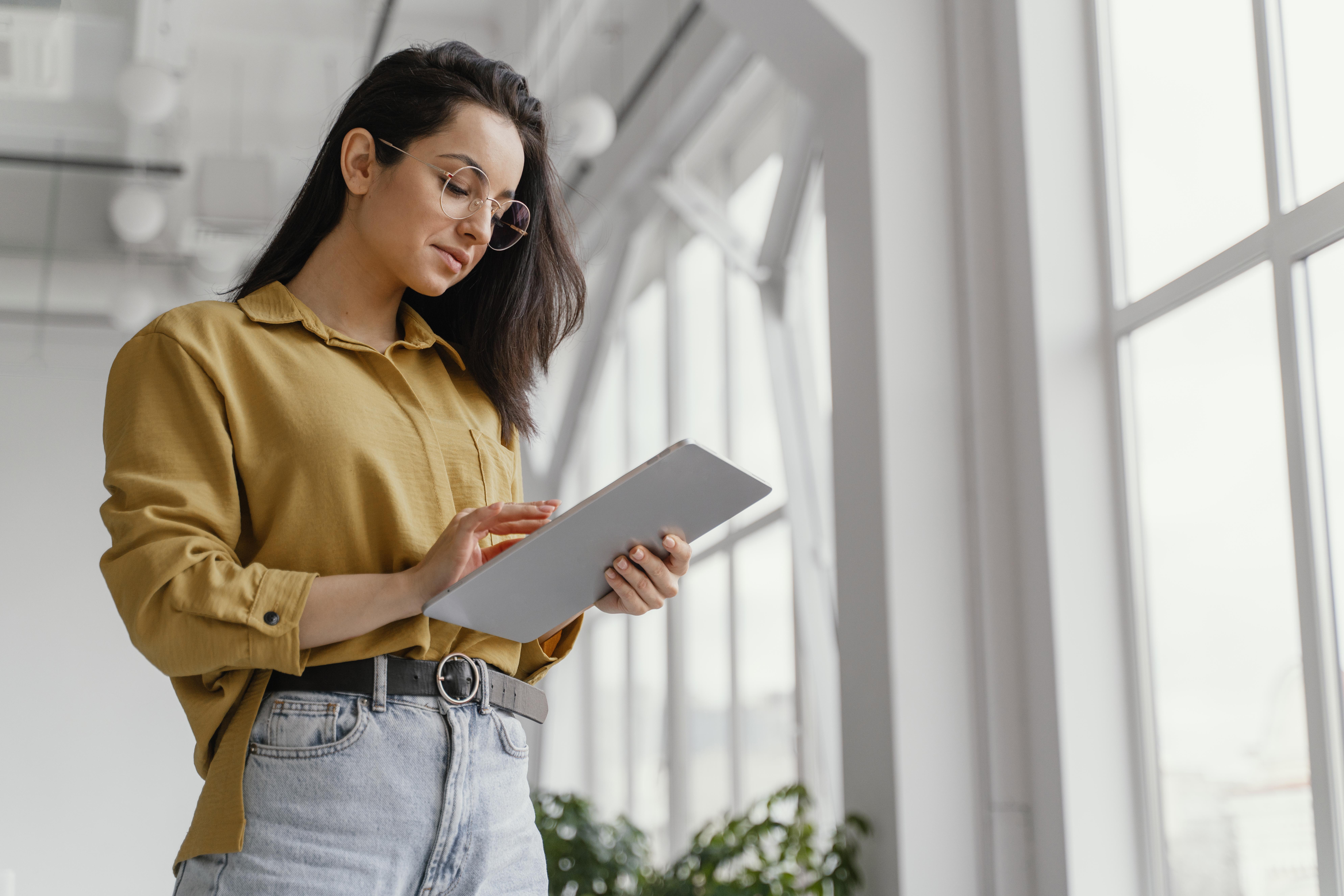 Mulher profissional usando um tablet em ambiente corporativo iluminado, analisando informações com atenção em um escritório moderno.